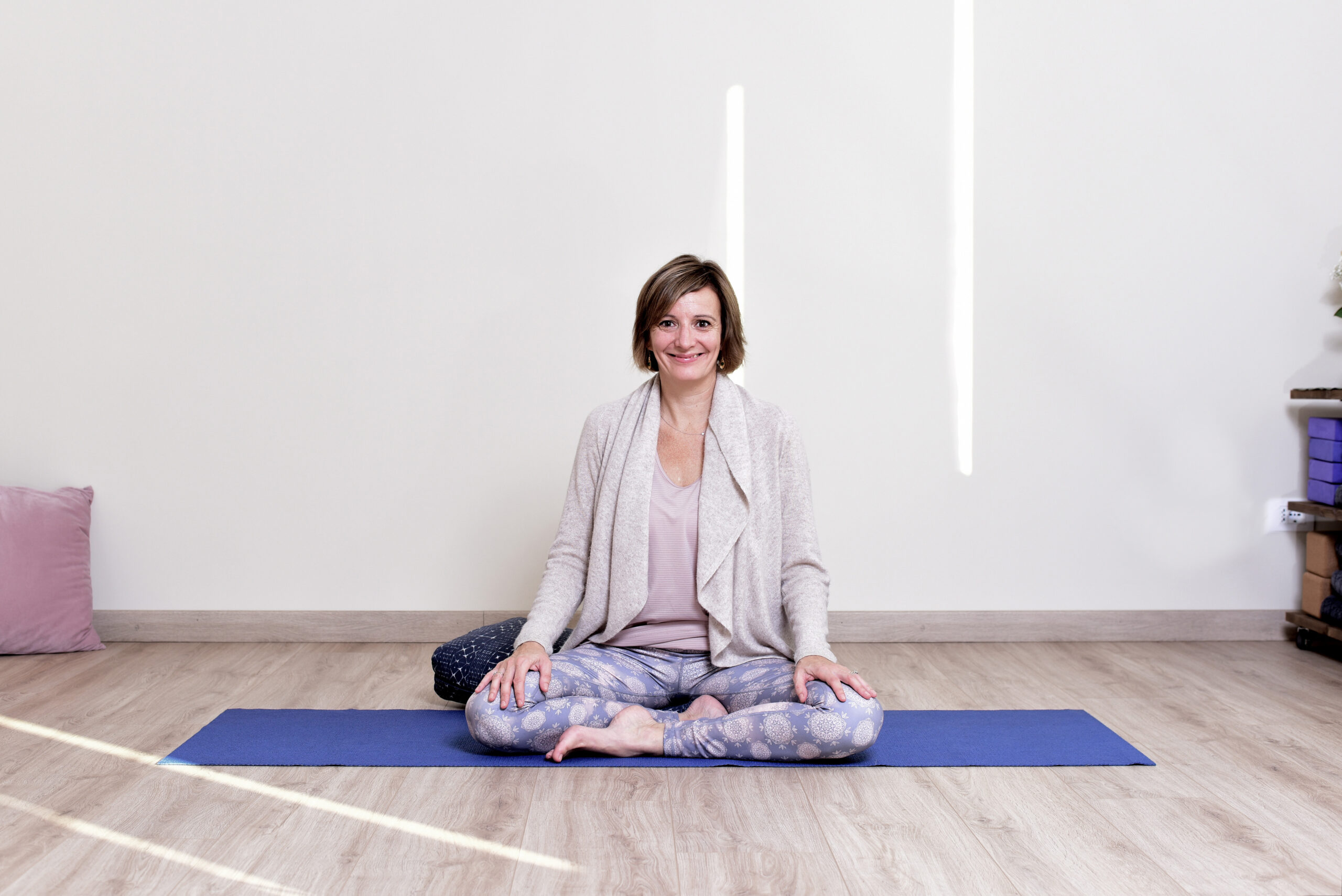 Audrey Favreau smiling, seated cross-legged on a blue mat in her studio