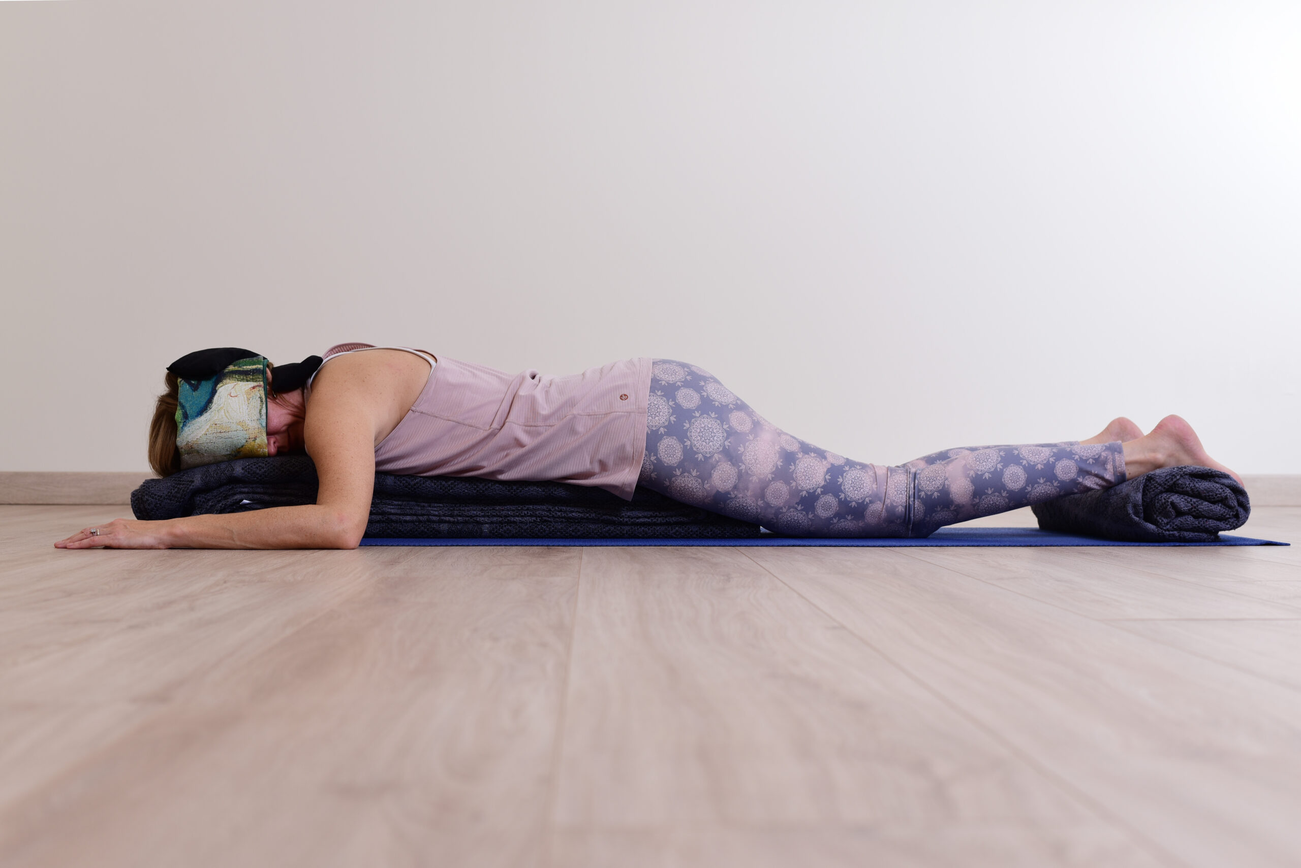 Audrey Favreau lying face down on a blue mat, her head turned to the side and resting on folded blankets, an eye pillow on her temple and her ankles supported by a rolled blanket