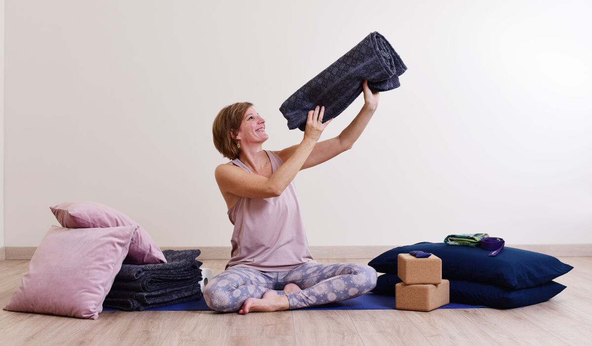 Audrey Favreau, sitting cross-legged and smiling, holds up a rolled blanket, surrounded by cushions, folded blankets, bolsters and yoga blocks